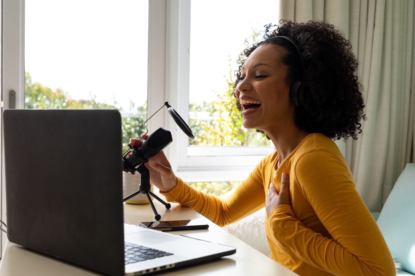 Happy biracial woman using headphones, laptop and microphone podcasting from home. Communication, technology, social media and domestic life, unaltered.